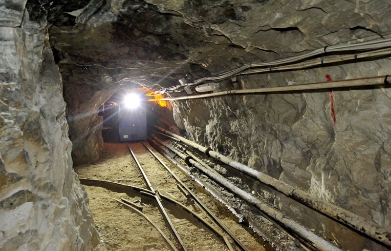 Galleria mineraria con binari e tubazioni illuminata dal trenino in arrivo.