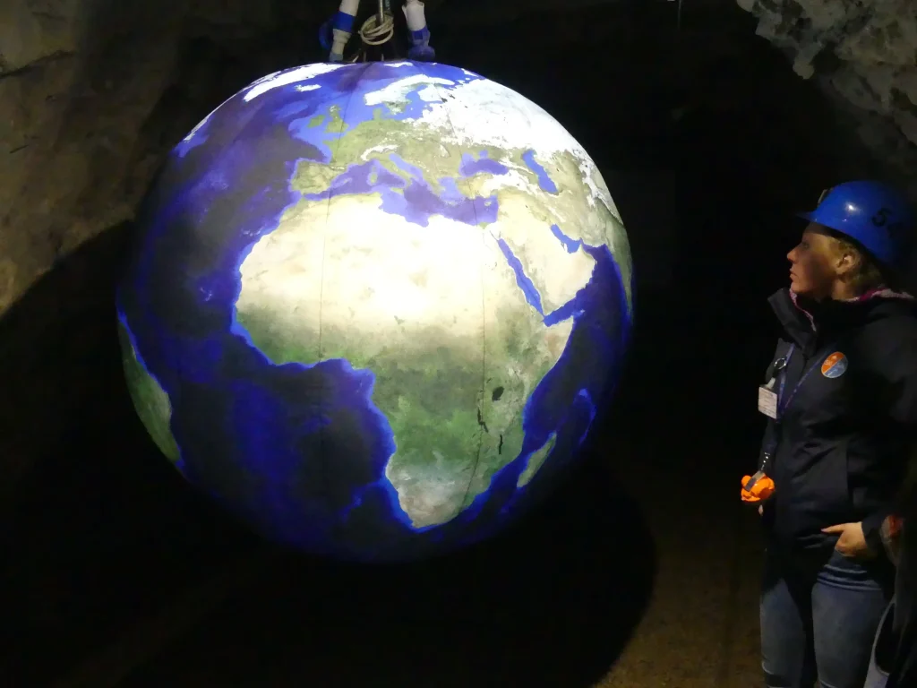 Mappamondo con immagine satellitare della Terra sospeso all'interno di una galleria mineraria, osservato da una visitatrice con casco.